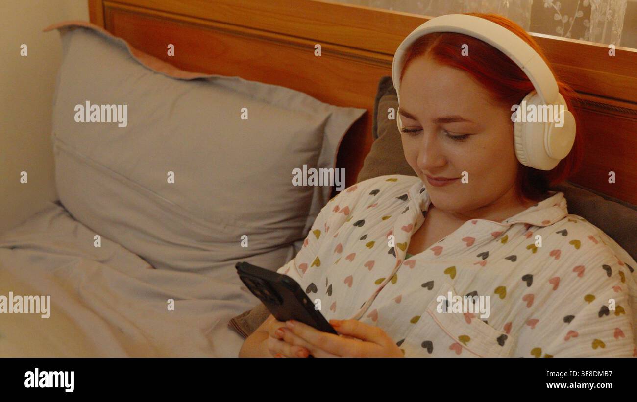 Giovane donna con le cuffie che usa lo smartphone a letto Foto Stock