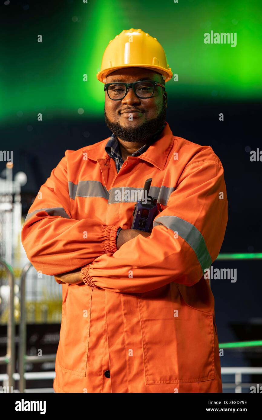 Ritratto di un allegro ingegnere di piattaforme offshore che supervisiona le operazioni durante l'aurora boreale. Jolly, esperto di carri di perforazione afroamericani, supervisiona le attività di produzione sul ponte e sullo sfondo dell'aurora boreale Foto Stock