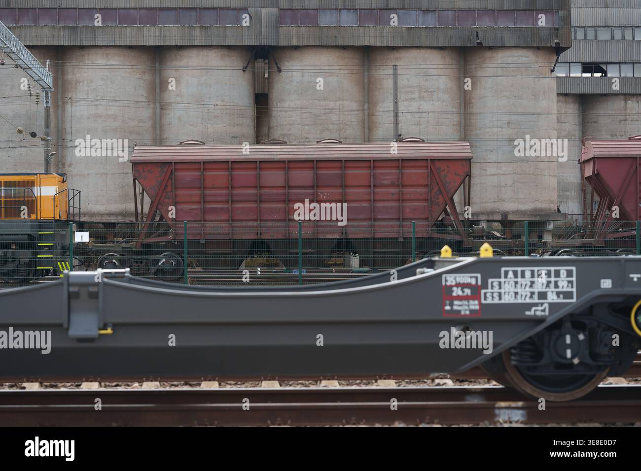 Carro tramoggia marrone per merci sfuse parcheggiato in un sito industriale ferroviario con silos in calcestruzzo sullo sfondo, trasporto merci su rotaia pesante e Grain st Foto Stock