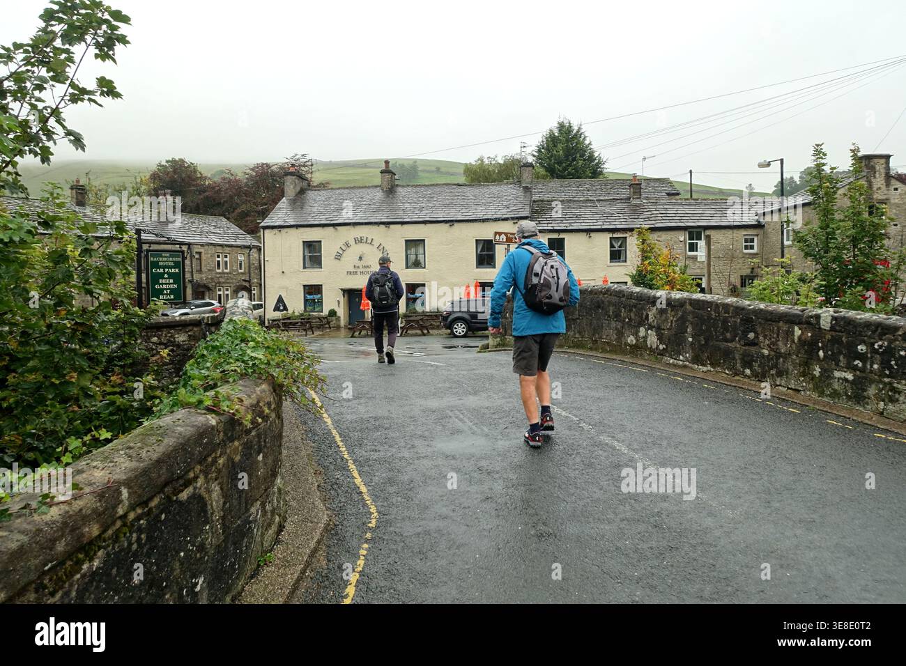 Due uomini anziani (escursionisti) camminando sul ponte fino al Blue Bell Inn, Kettlewell, Wharfedale nello Yorkshire Dales National Park, Inghilterra, Regno Unito. Foto Stock