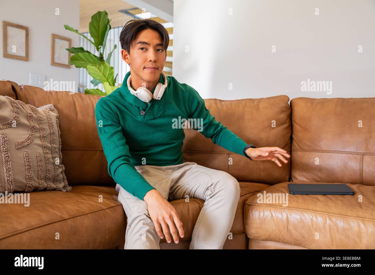 Uomo asiatico seduto su un divano sezionale in pelle abbronzata con cuffie bianche, in cerca di un tablet Foto Stock
