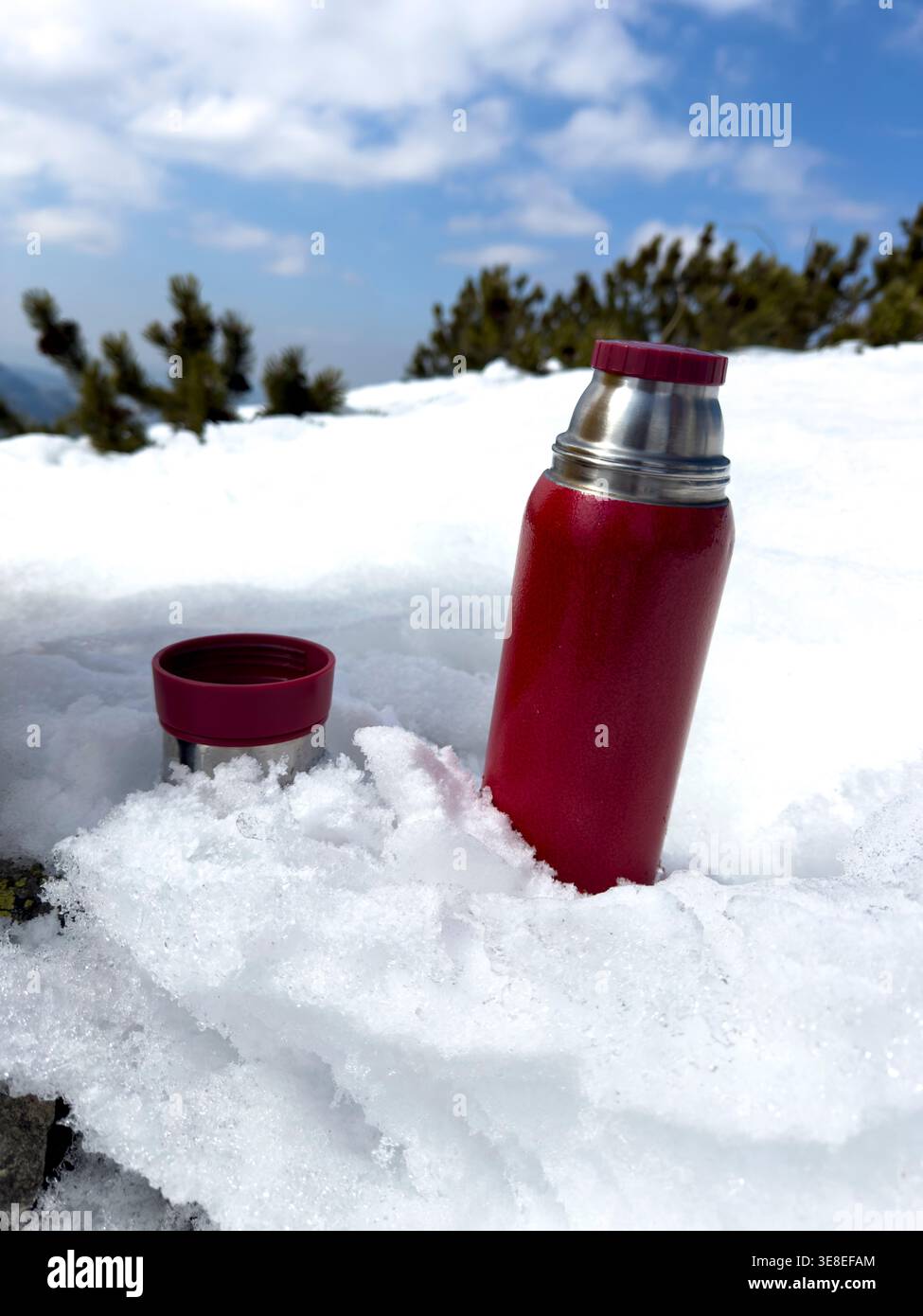 La bottiglia rossa thermos sulla neve sul sentiero di montagna presenta l'avventura invernale di viaggio idratazione all'aperto attrezzatura da campeggio riutilizzabile per bere da trekking touri - Immagine stock catturata con smartphone