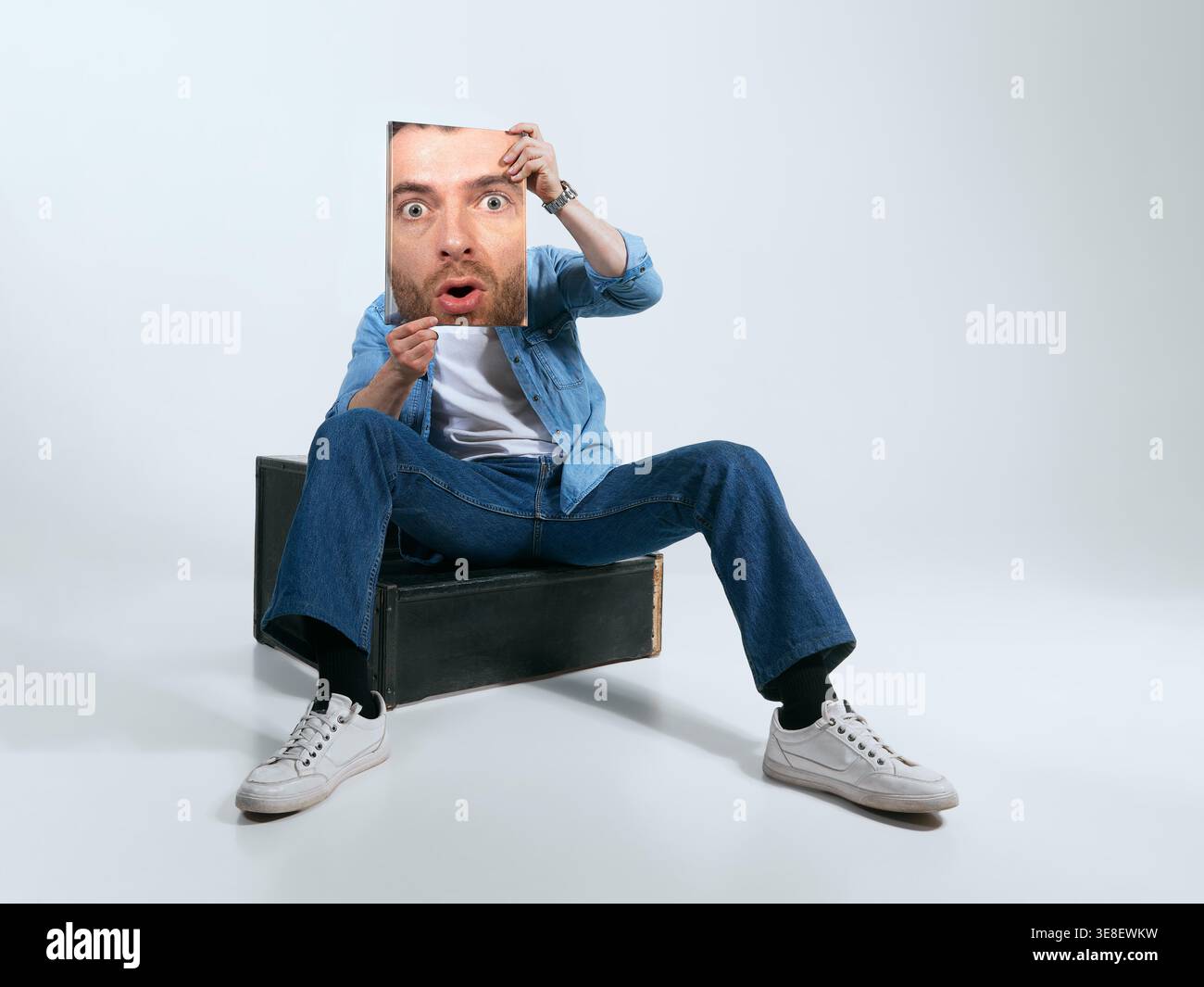 Uomo con abito in denim seduto su una scatola che regge il ritratto di uno studio di stampa del volto sorpreso. Foto Stock