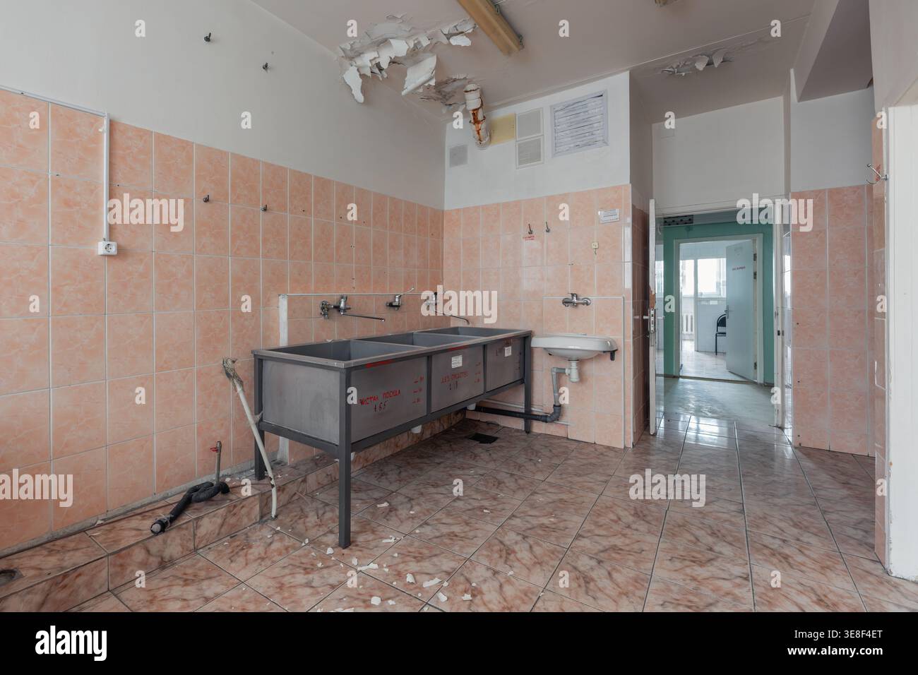 Stanza di servizio in un ospedale di maternità sovietico abbandonato con lavandini in acciaio, soffitto danneggiato, tubazioni esposte e pareti piastrellate. Quelli trascurati Foto Stock