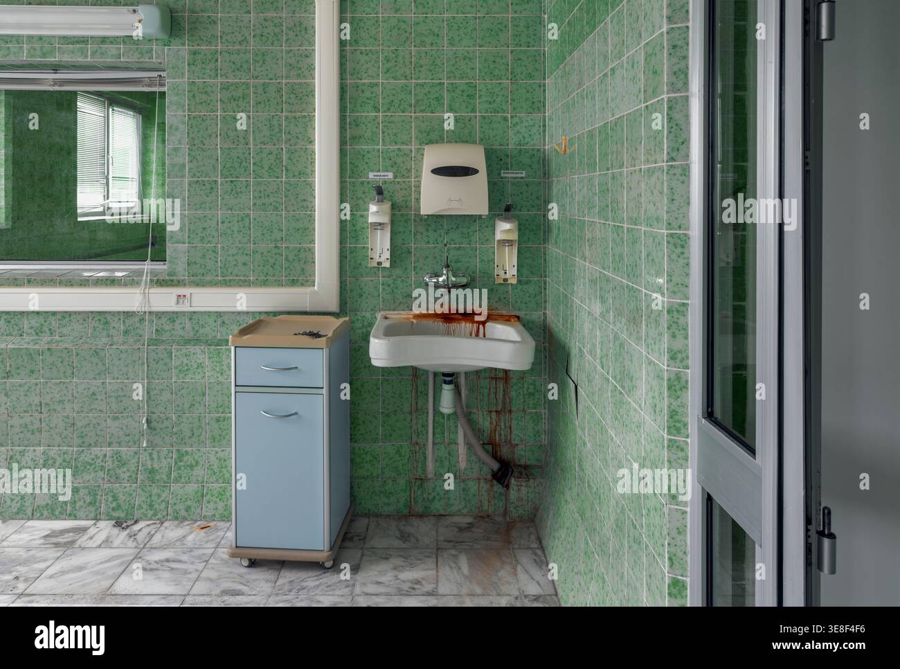 Lavello macchiato di ruggine in un ospedale di maternità sovietico abbandonato, circondato da piastrelle verdi e dispenser vuoti. L'interno medico trascurato riflette d Foto Stock