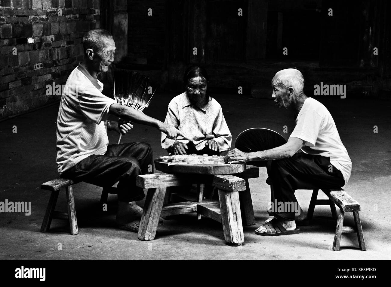 Due uomini anziani che giocano a un gioco da tavolo, con una terza persona che guarda, in una stanza poco illuminata fotografie in bianco e nero che catturano un momento di svago Foto Stock