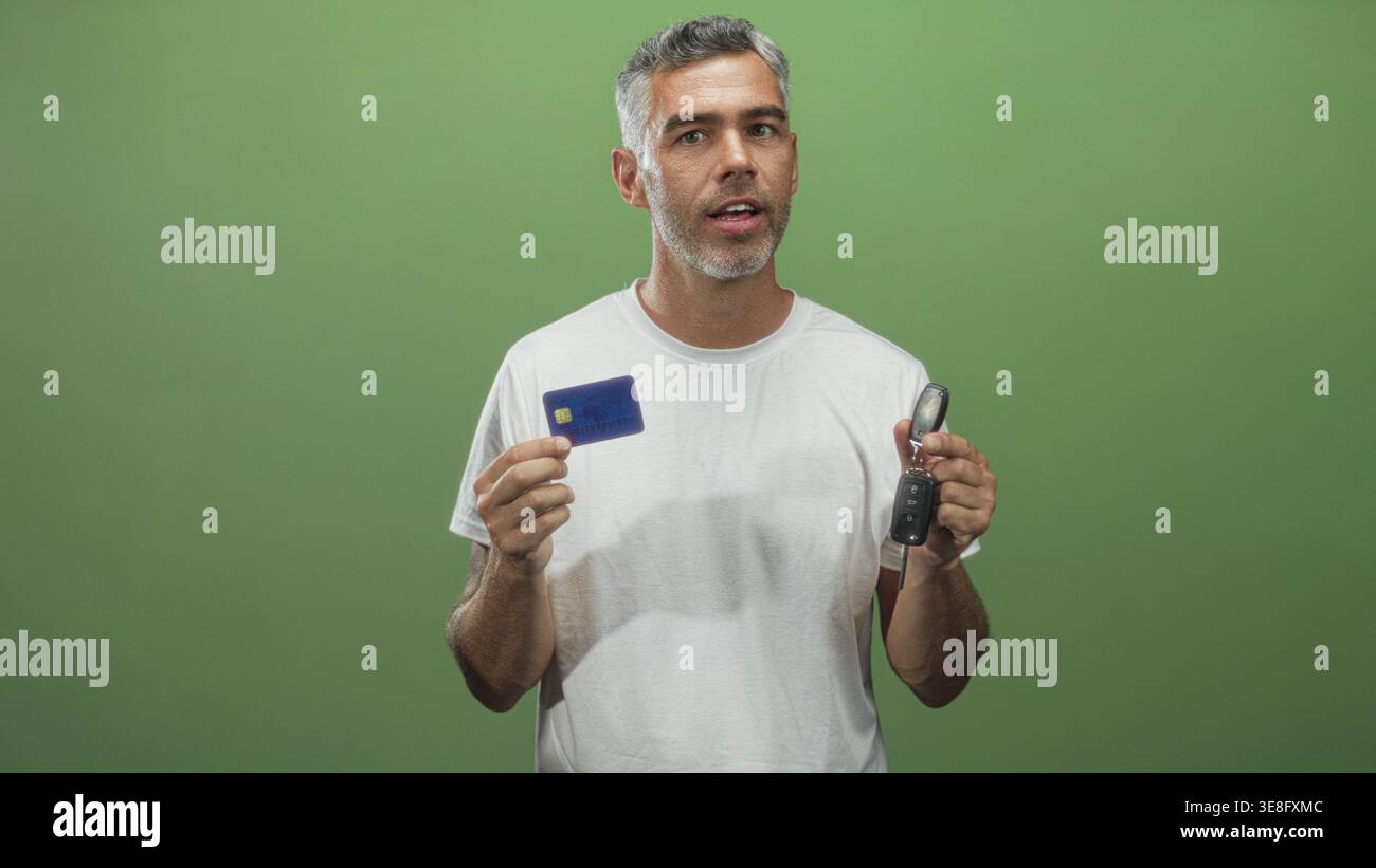 Uomo di mezza età con i capelli grigi tiene una carta di credito e le chiavi dell'auto in una mano, l'altra mano vicino al petto, gli occhi chiusi, indossa una t shirt bianca in studio; fi Foto Stock