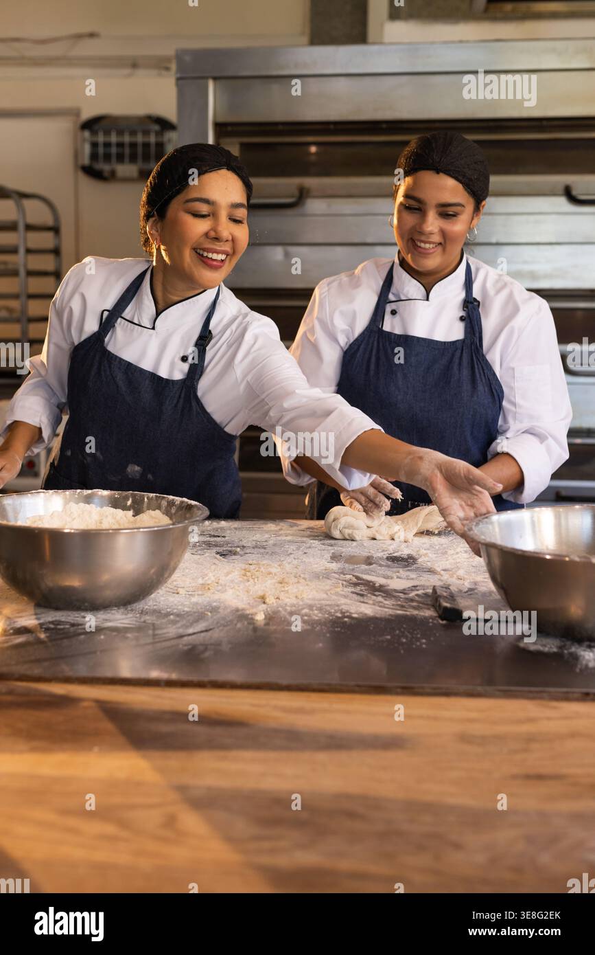 Due cuoche femminili in giacche da chef, grembiuli che impastano, si avvicinano a ciotole di metallo a tavola infarinata, forni Foto Stock