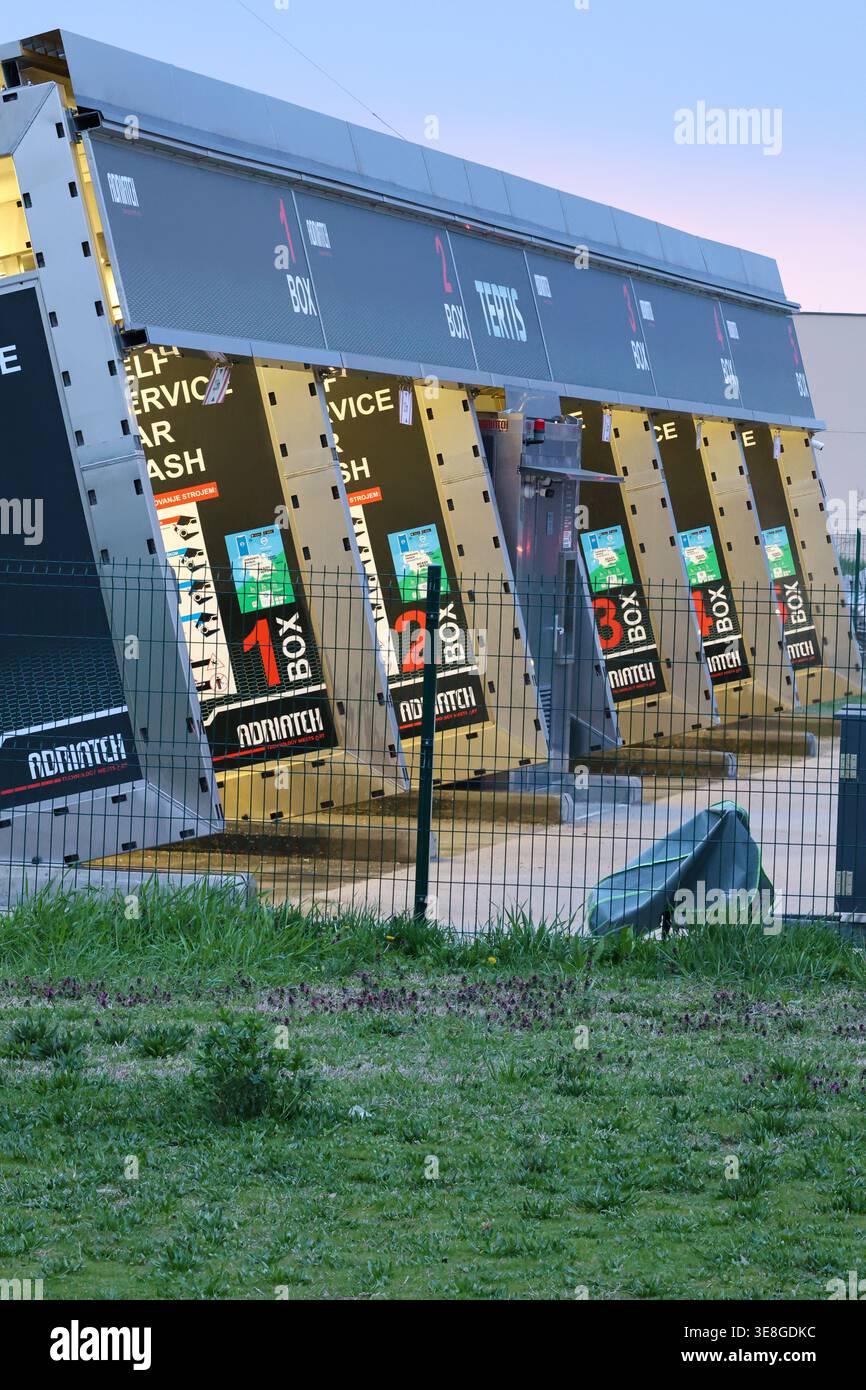 Vinkovci, Croazia - 22 marzo 2026: Fila di banchi di autolavaggio self-service illuminati al crepuscolo, con stazioni di lavaggio automatiche numerate, attrezzature e recinzioni W Foto Stock