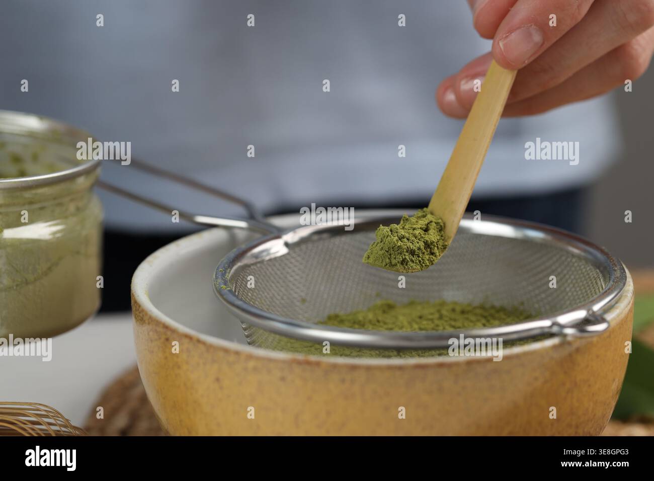 Donna setacciando polvere di matcha nel recipiente al tavolo bianco, primo piano Foto Stock