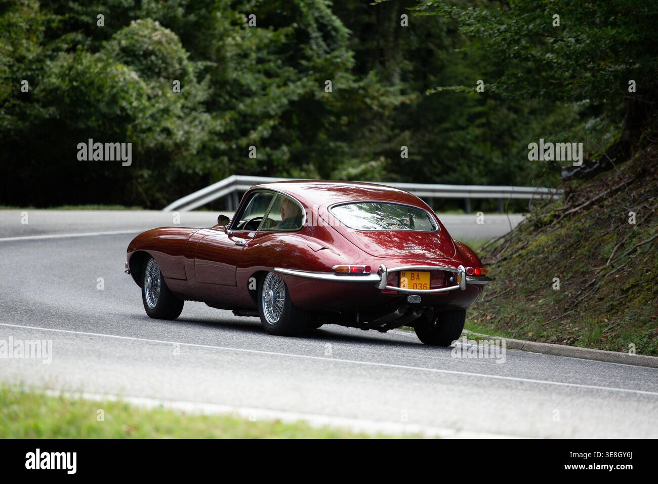 Vista posteriore di una Jaguar e-Type Sportscar rossa profonda classica che guida su una Winding Mountain Road Foto Stock