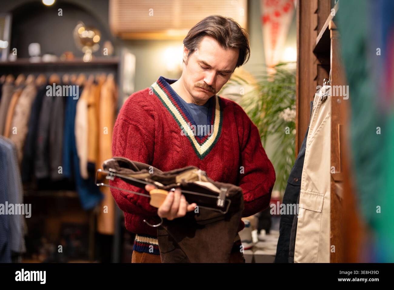 Un cliente alla moda attento all'uomo hipster che sceglie pantaloni di velluto alla moda in un negozio vintage di seconda mano. Foto Stock
