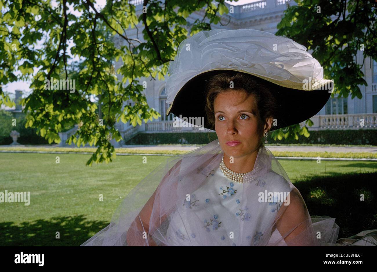 Fotografia storica a colori della signora Hutton seduta all'aperto alla Marble House, con un elegante cappello e un abbigliamento formale con perle, adagiata contro i giardini paesaggistici e la grandiosa architettura della tenuta di Vanderbilt, fotografata da Toni Frissell. Immagine della moda e dello stile di vita della metà del XX secolo che cattura ricchezza, raffinatezza e cultura della società. Foto Stock