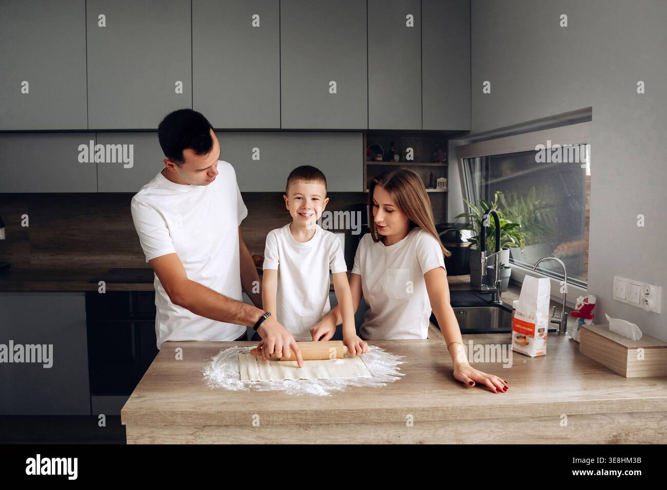 La famiglia si riunisce in cucina per preparare l'impasto sul piano di lavoro mentre il bambino aiuta a arrotolare. La farina viene sparsa in giro mentre condividono i momenti durante la cottura dell'aria condizionata Foto Stock