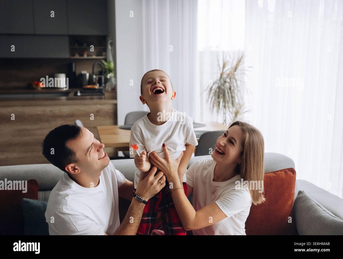 Un bambino ride gioiosamente mentre viene sollevato da due adulti in un soggiorno. Lo spazio ha un design moderno, con piante e luce solare che passano attraverso Foto Stock