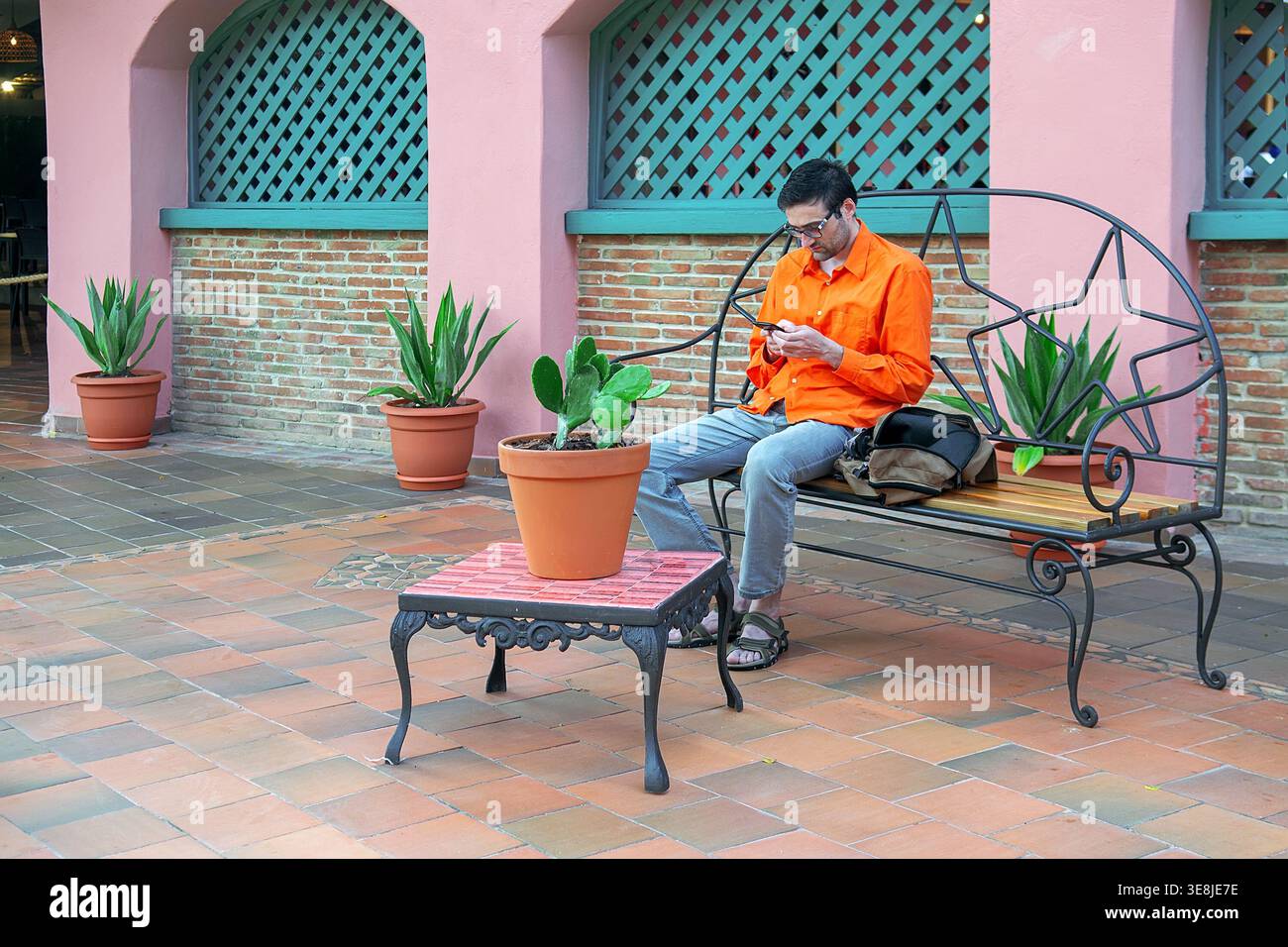 L'uomo hipster con la camicia arancione studia il menu sullo smartphone mentre è seduto sulla panchina circondato da cactus in vaso sul patio piastrellato del ristorante messicano all-i. Foto Stock
