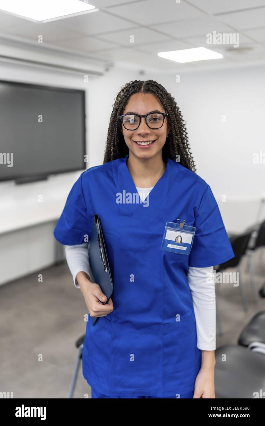 Giovane studentessa di sanità che indossa uniforme medica blu, occhiali, e un badge di identificazione, tenendo una cartella e sorridendo con orgoglio in un moderno luminoso Foto Stock
