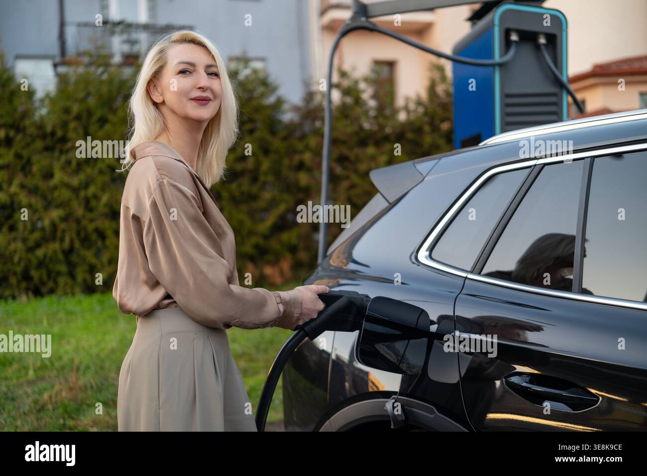 Il proprietario del veicolo elettrico collega il caricabatterie all'auto presso la stazione di ricarica esterna. Moderno ambiente residenziale con vegetazione ed edifici visibili sullo sfondo. Foto Stock