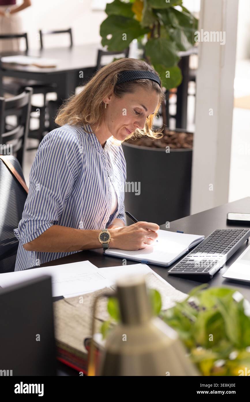 Donna adulta che scrive su un notebook aperto in un ufficio condiviso e indossa una camicia a righe con tastiera Foto Stock