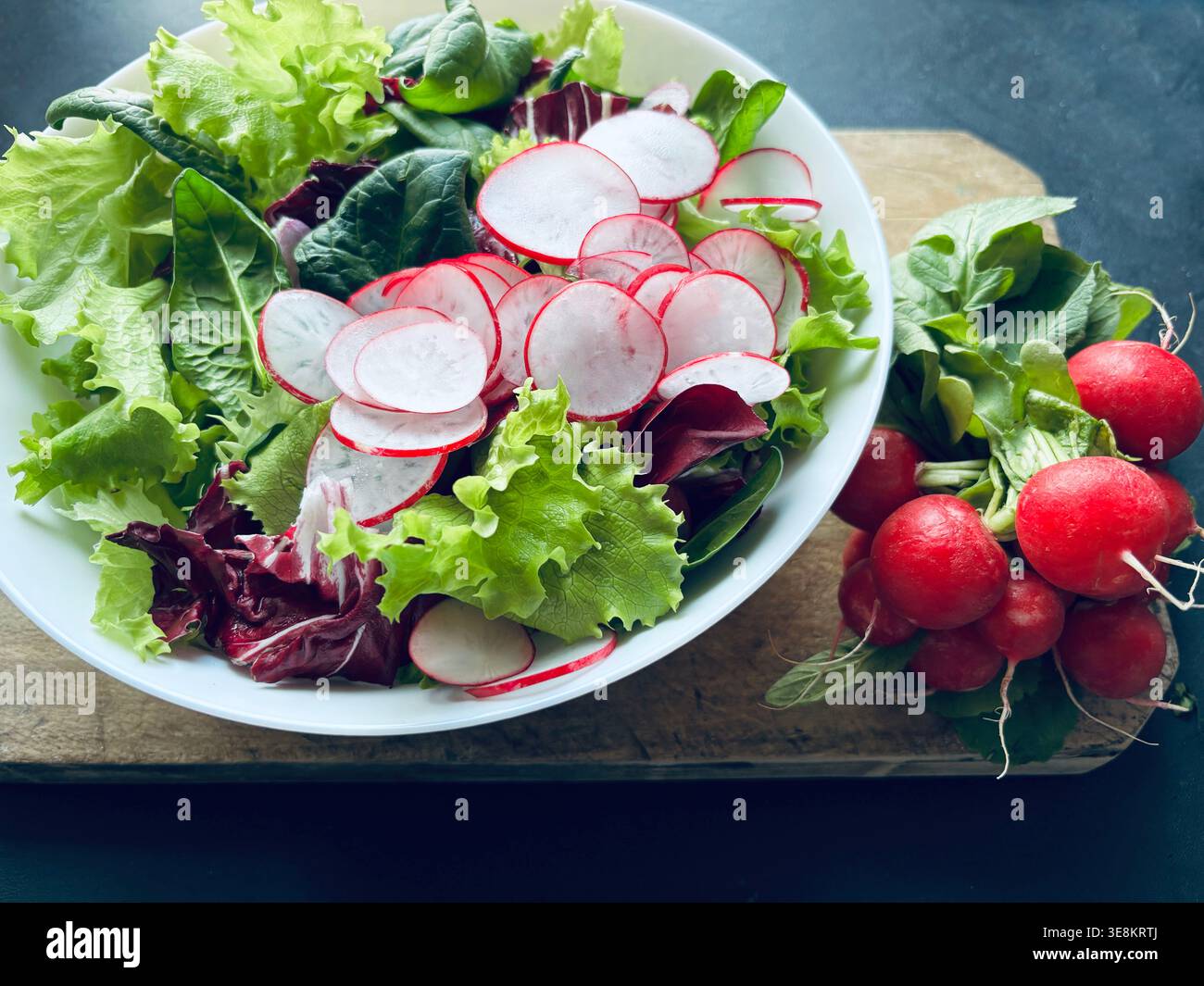 Insalata mista fresca con ravanelli tagliati a fette servita in una ciotola bianca su un rustico tavolo di legno. Un mazzo di raggi interi sono serviti accanto. Mangiare sano Foto Stock