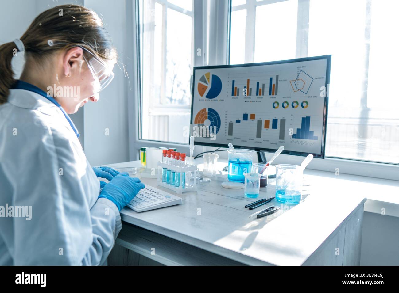 Giovane ricercatore che indossa occhiali protettivi, i guanti lavorano sulla tastiera in un laboratorio illuminato dal sole. Analisi dei dati, ricerca scientifica, esperimenti di laboratorio sono evidenziati in un ambiente pulito e ad alta tecnologia Foto Stock