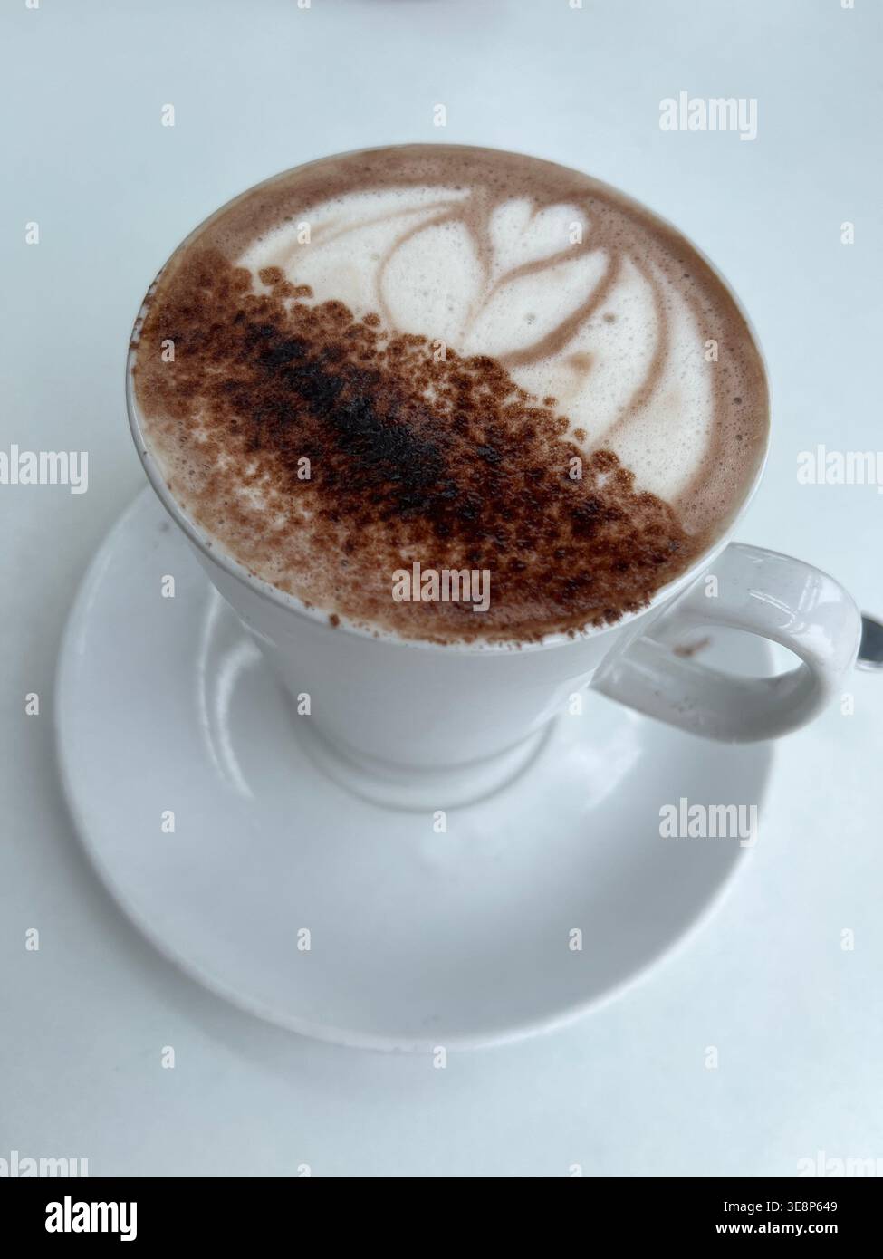 Latte appena fatto con intricata schiuma art in una tazza e un piattino in ceramica bianca, servito su un tavolo da caffè con luce naturale soffusa e messa a fuoco selettiva. - Immagine stock catturata con smartphone