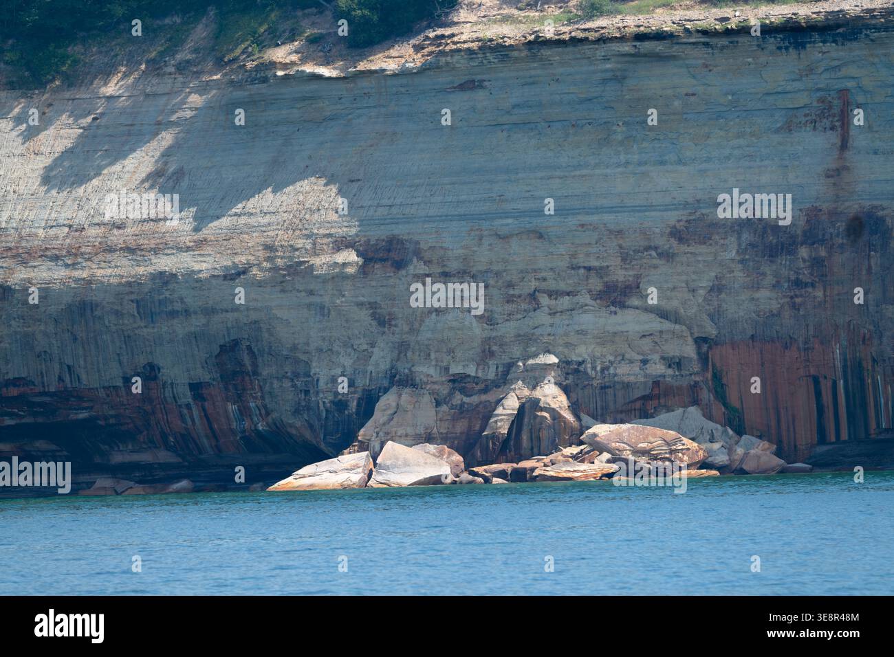 La luce del sole proietta ombre sulle torreggianti scogliere che si innalzano sopra acque tranquille. Le formazioni rocciose mostrano gli strati della terra mentre le pietre lisce d Foto Stock