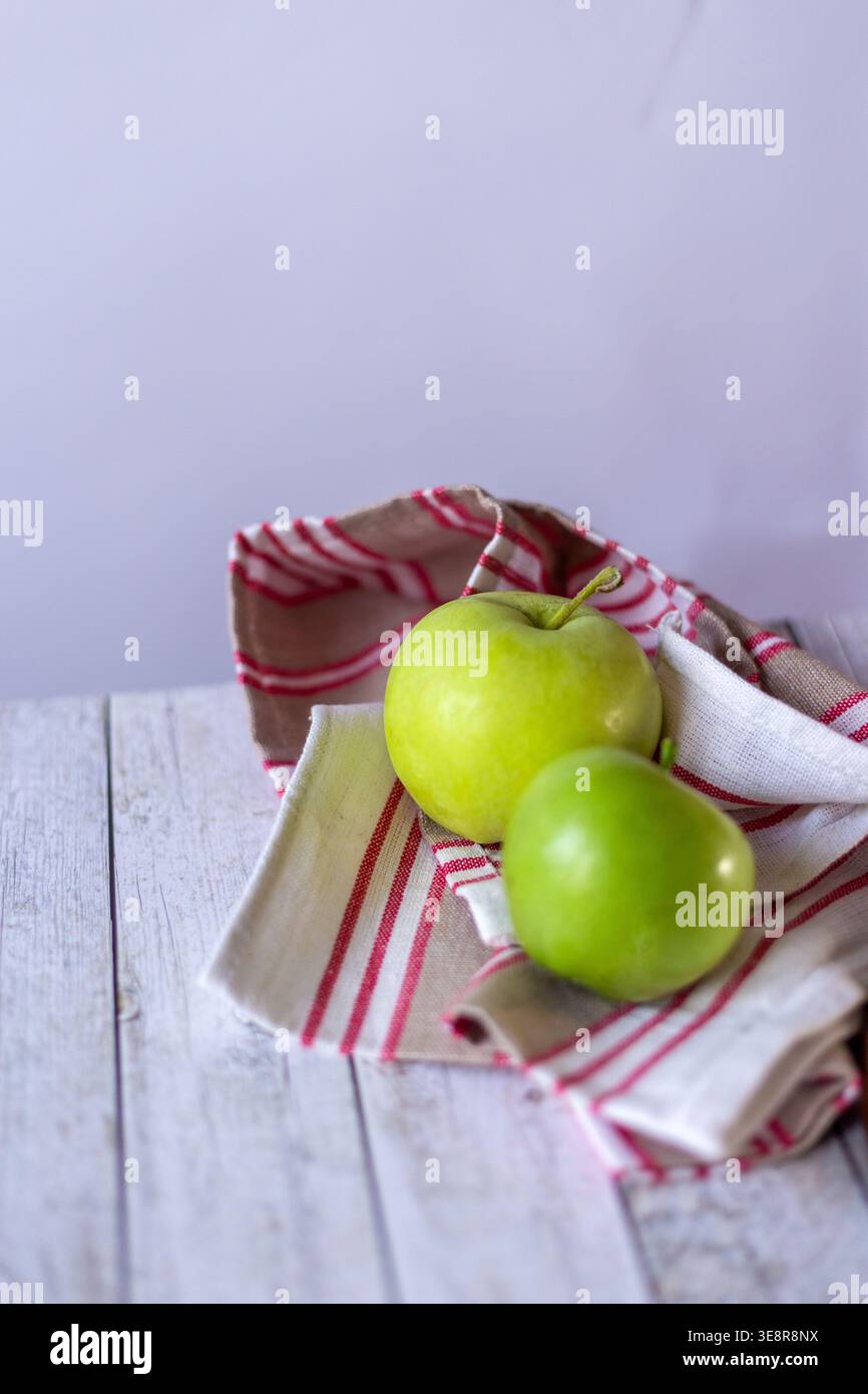 Due mele verdi poggiano su un panno a righe rosso e beige collocato su un tavolo di legno chiaro in cucina. La luce naturale illumina la scena. Foto Stock