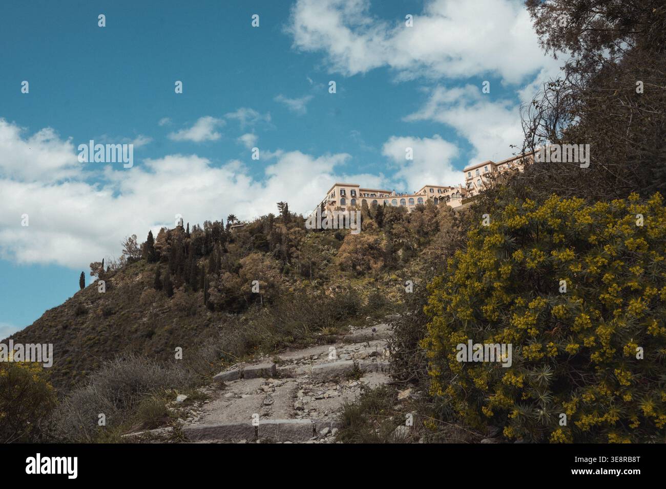 Vista delle antiche scale di pietra che si arrampicano verso un villaggio in cima a una collina tra lussureggianti alberi verdi sullo sfondo di cielo blu e nuvole bianche, Taormina e siti Foto Stock