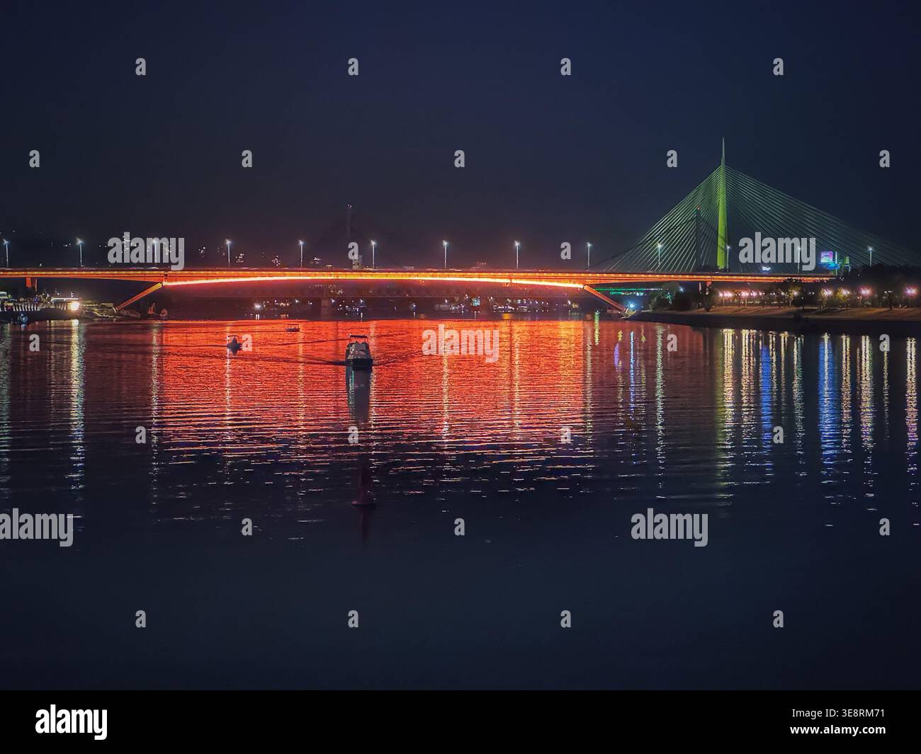 Vista notturna del ponte Gazela sul fiume Sava a Belgrado, in Serbia, con le luci della città che si riflettono sull'acqua. Paesaggio urbano e infrastrutture moderne - Immagine stock catturata con smartphone