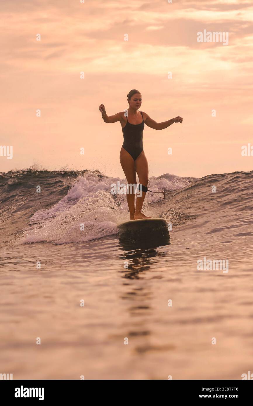 Donna che naviga sotto il cielo rosa al tramonto, silhouette rilassata su Longboard con retroilluminazione morbida e delicata Foto Stock