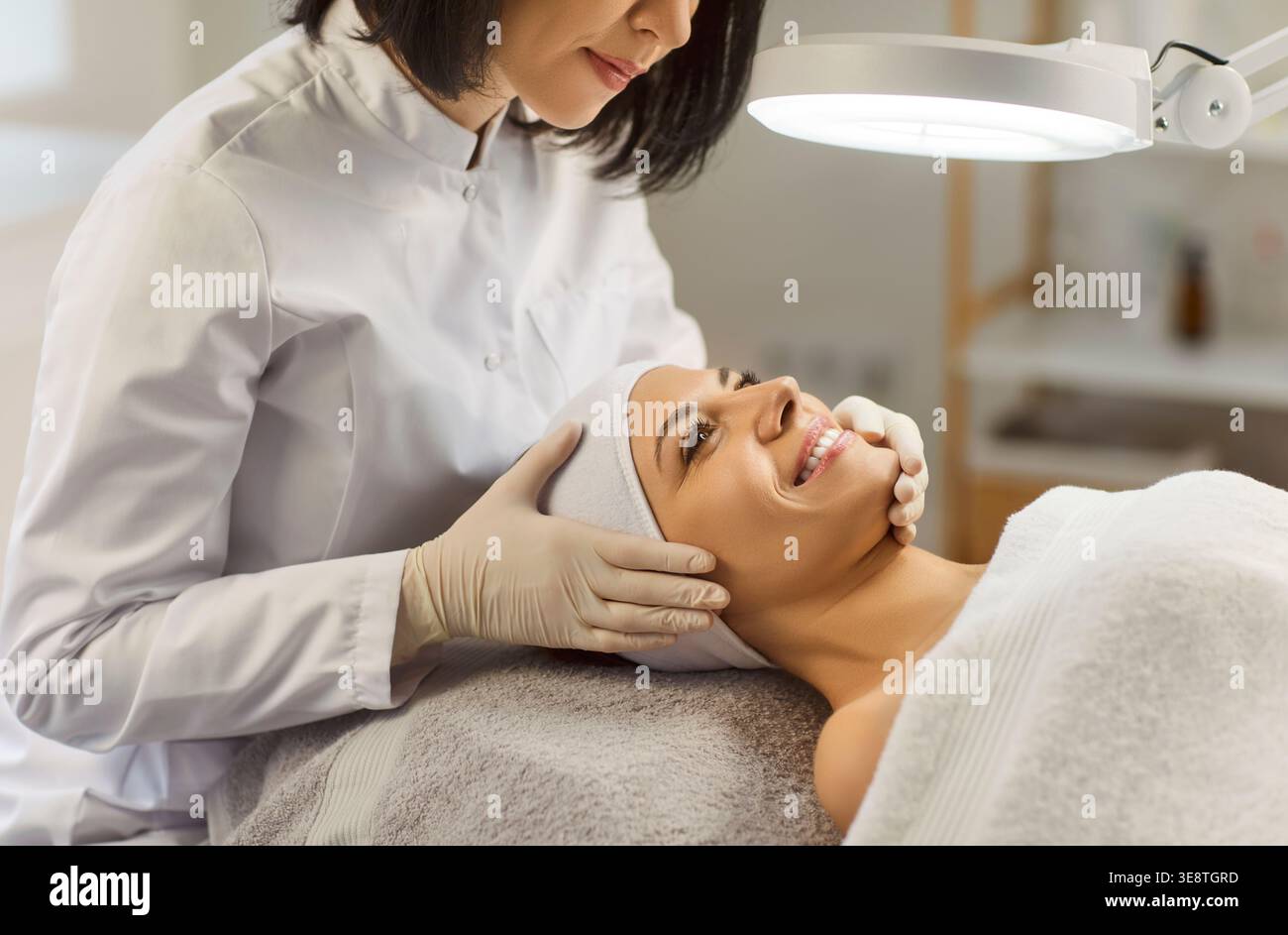 Cosmetologa professionista che esamina la pelle del viso del cliente, dirige la lampada per una buona visione Foto Stock