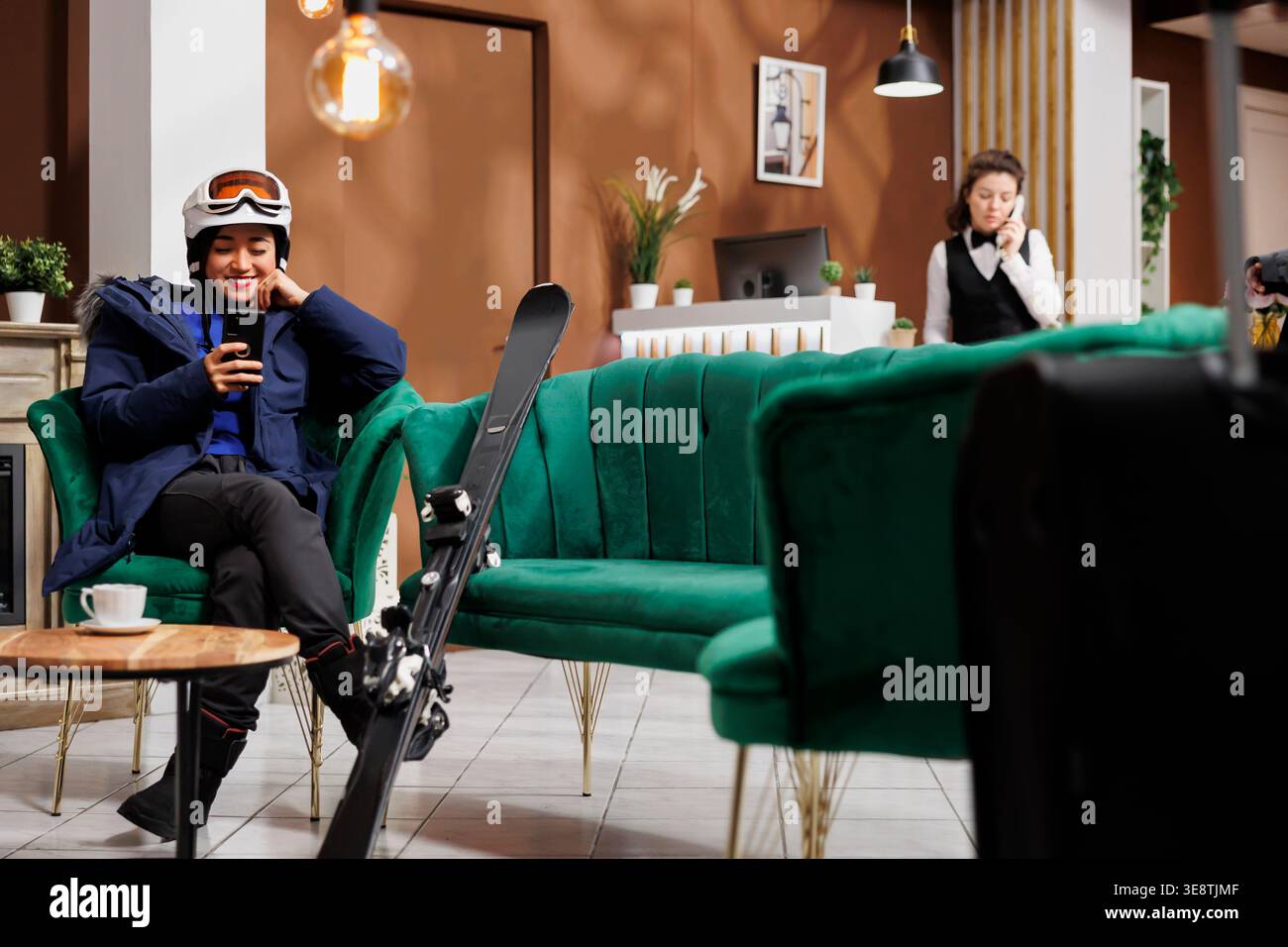 Signora asiatica entusiasta che usa il suo telefono cellulare e aspetta nella hall dell'hotel per prenotare la procedura per le vacanze invernali. Viaggiatore in attrezzatura per sport invernali con il suo smartphone nella zona lounge della stazione sciistica. Foto Stock