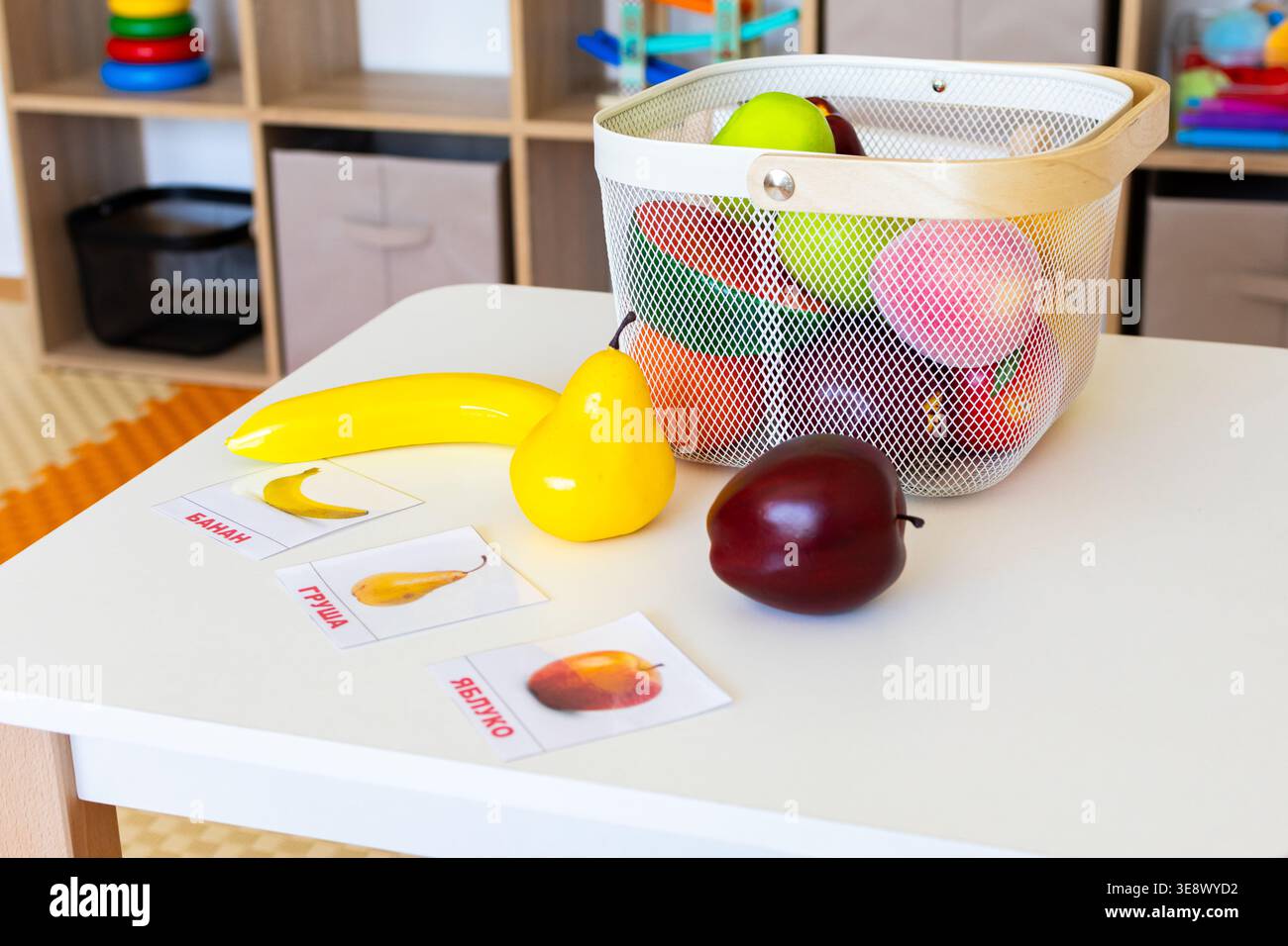 Il cestino di frutta giocattolo si trova su una scrivania per bambini con banana di mele e prugna accanto alle carte etichettate, creando una postazione giocosa per la selezione dei nomi e dei colori Foto Stock