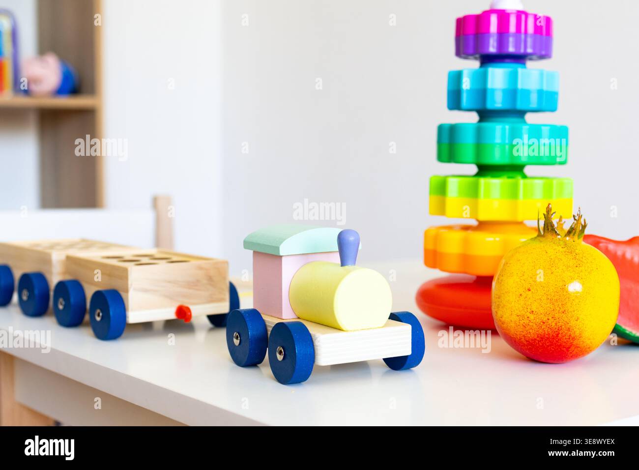 Il trenino giocattolo in legno con ruote blu rotola su un tavolo per bambini accanto a una torre di impilamento arcobaleno e gioca a frutta ispirando storie di trasporto fantasiose Foto Stock