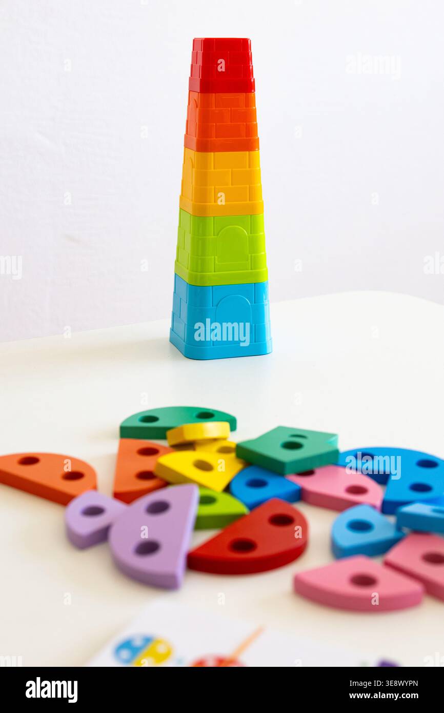 Un'alta torre arcobaleno si erge su un tavolo bianco mentre nelle vicinanze si spargono pezzi di legno grossi. I colori vivaci invitano i più piccoli a ordinare pila e immaginarla Foto Stock