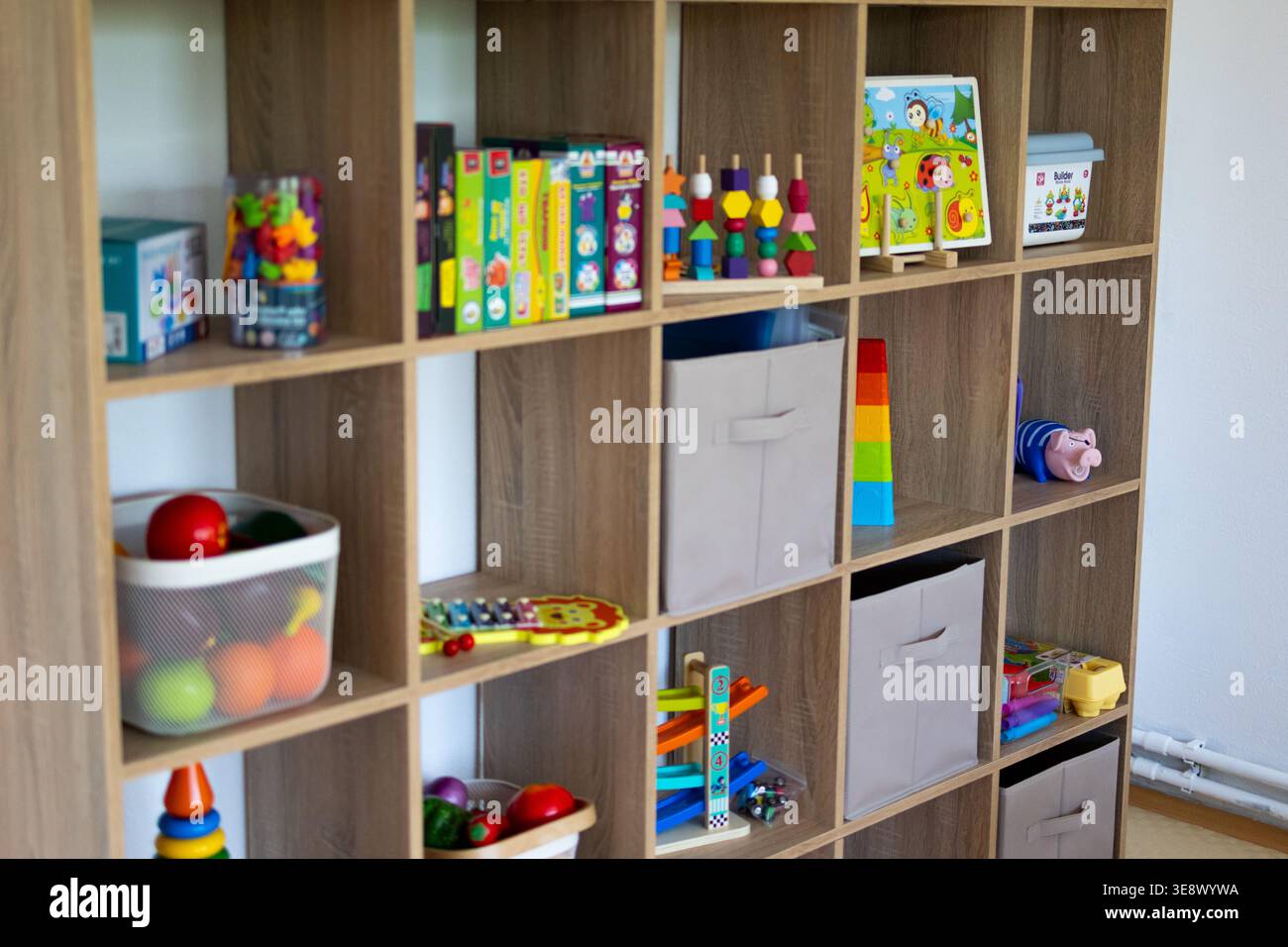 Ampia scaffalatura cubica piena di giocattoli didattici, puzzle e bidoni in tessuto in una luminosa sala giochi. Gli scomparti organizzati mostrano oggetti colorati Foto Stock
