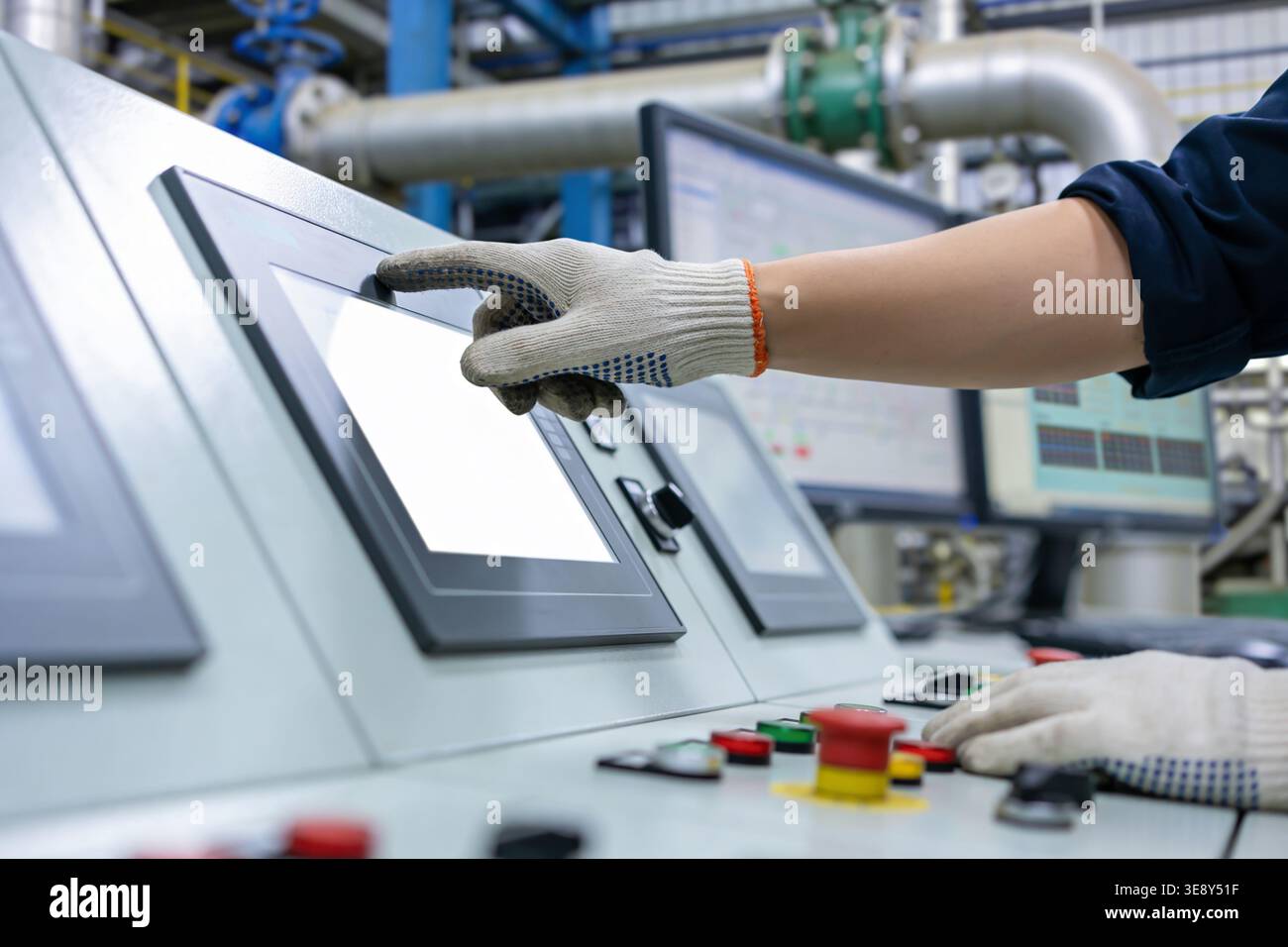 Un operatore che indossa guanti protettivi tocca uno schermo vuoto su un sofisticato sistema di controllo in fabbrica, mentre i monitor visualizzano i dati in Foto Stock