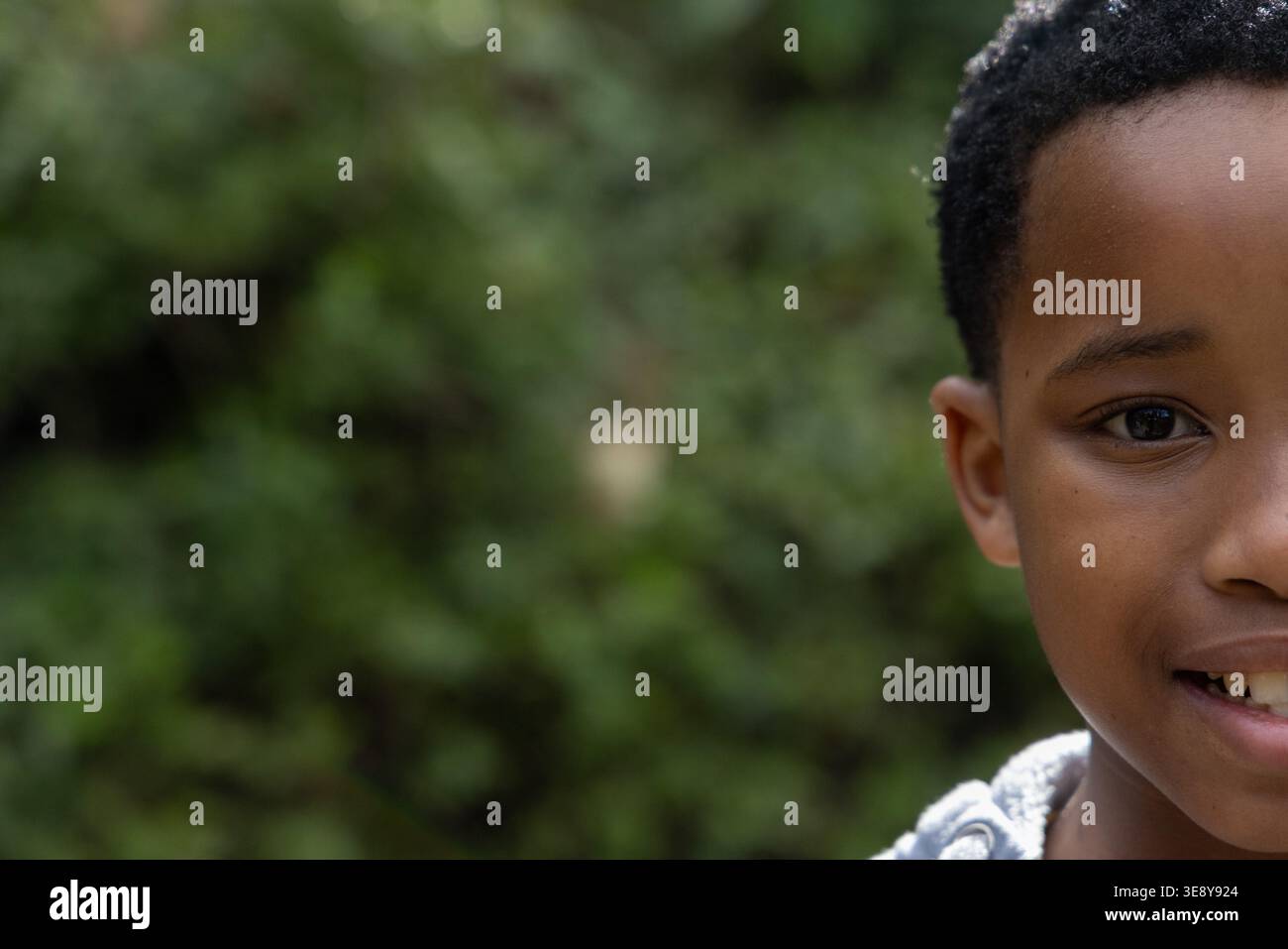 Bambino afroamericano che sorride, guarda la macchina fotografica nel parco con cappuccio grigio, copia spazio. Ritratto, esterno, natura, vegetazione, bokeh, illuminazione, occhio Foto Stock