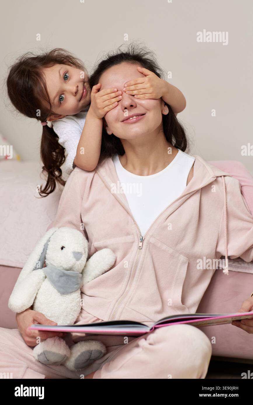 Madre e figlia giocano insieme, bambini che coprono gli occhi della mamma da dietro, condividendo momenti felici a casa Foto Stock