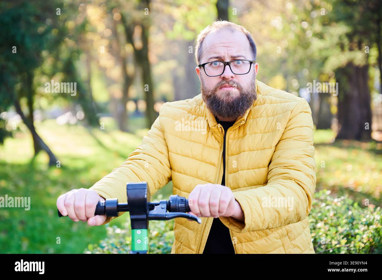Uomo caucasico in scooter esitante prima di guidare, espressione preoccupata e leggero appoggiarsi alla schiena tenendo il manubrio vicino agli alberi del parco, giacca gialla e occhiali per creare un'atmosfera cauto Foto Stock