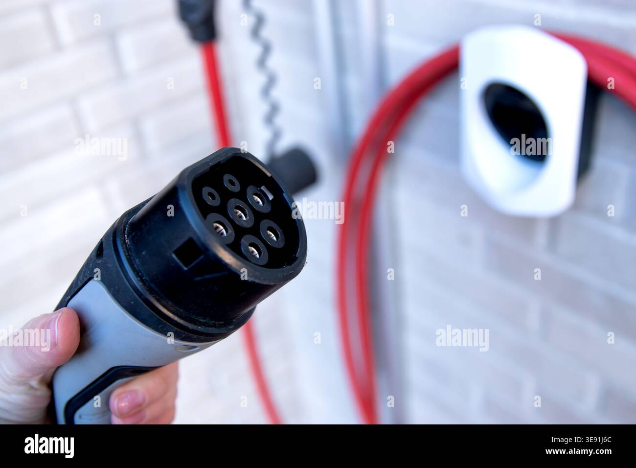 Connettore di ricarica per veicoli elettrici tenuto in mano, con una stazione di ricarica montata a parete e cavi rossi visibili sullo sfondo Foto Stock