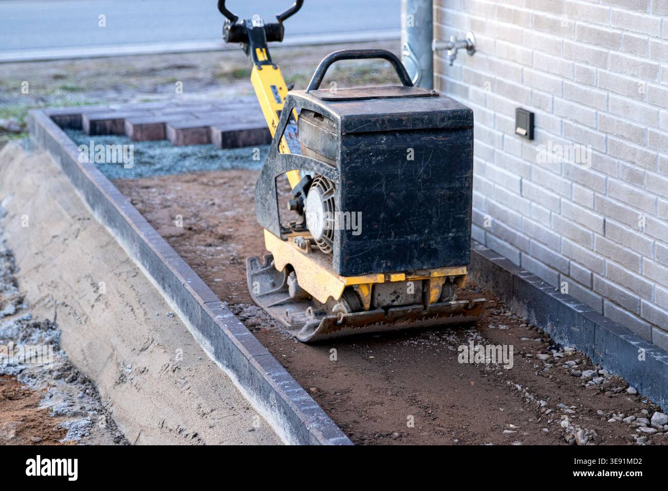 Compattatore a piastre per impieghi gravosi in cantiere che comprime il terreno vicino a una parete di mattoni, con ghiaia e tavole di legno visibili sullo sfondo Foto Stock