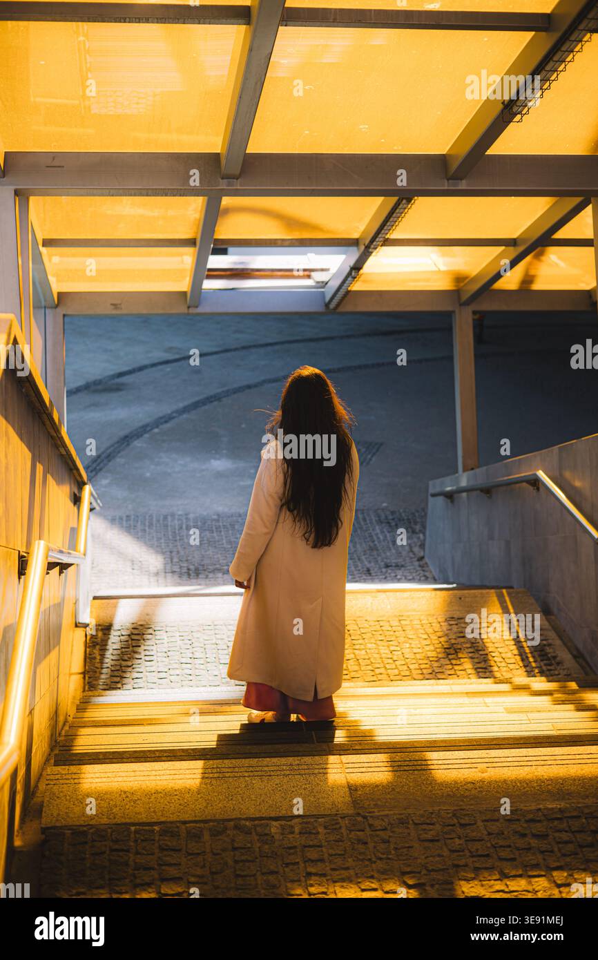 Una giovane donna con un cappotto beige che cammina giù per le scale della città al tramonto Foto Stock