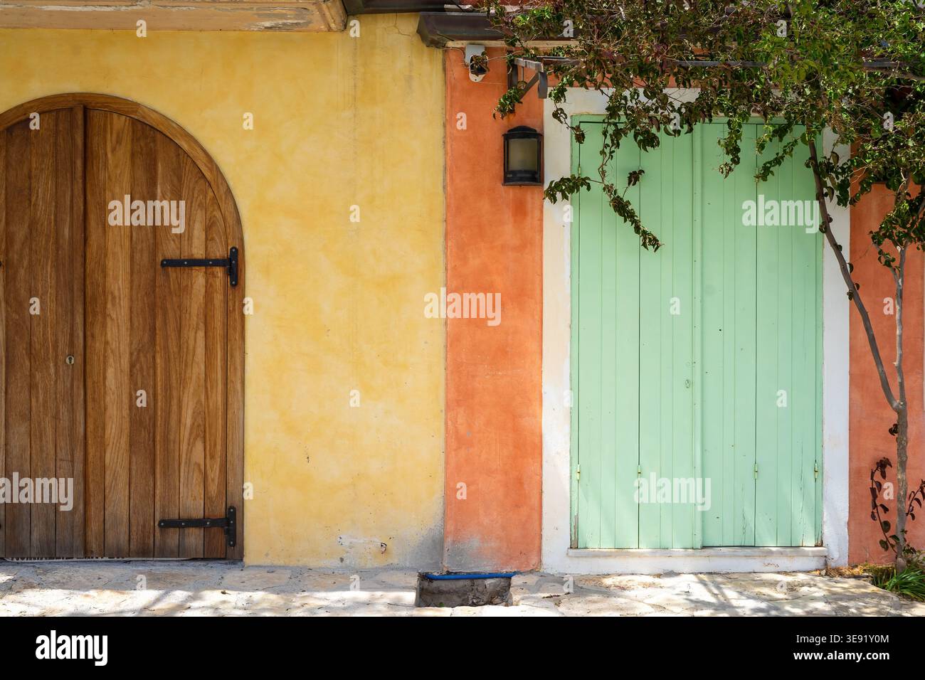 Due porte a contrasto, una in legno ad arco e una tapparella verde menta, adagiate su pareti in stucco giallo e arancione con fogliame. Foto Stock