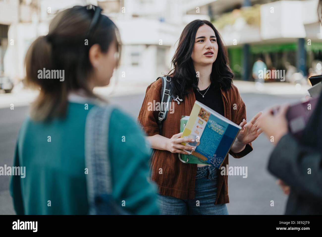 Studenti che parlano con gli amici nel campus tenendo un libro di testo e uno zaino in un ambiente urbano Foto Stock