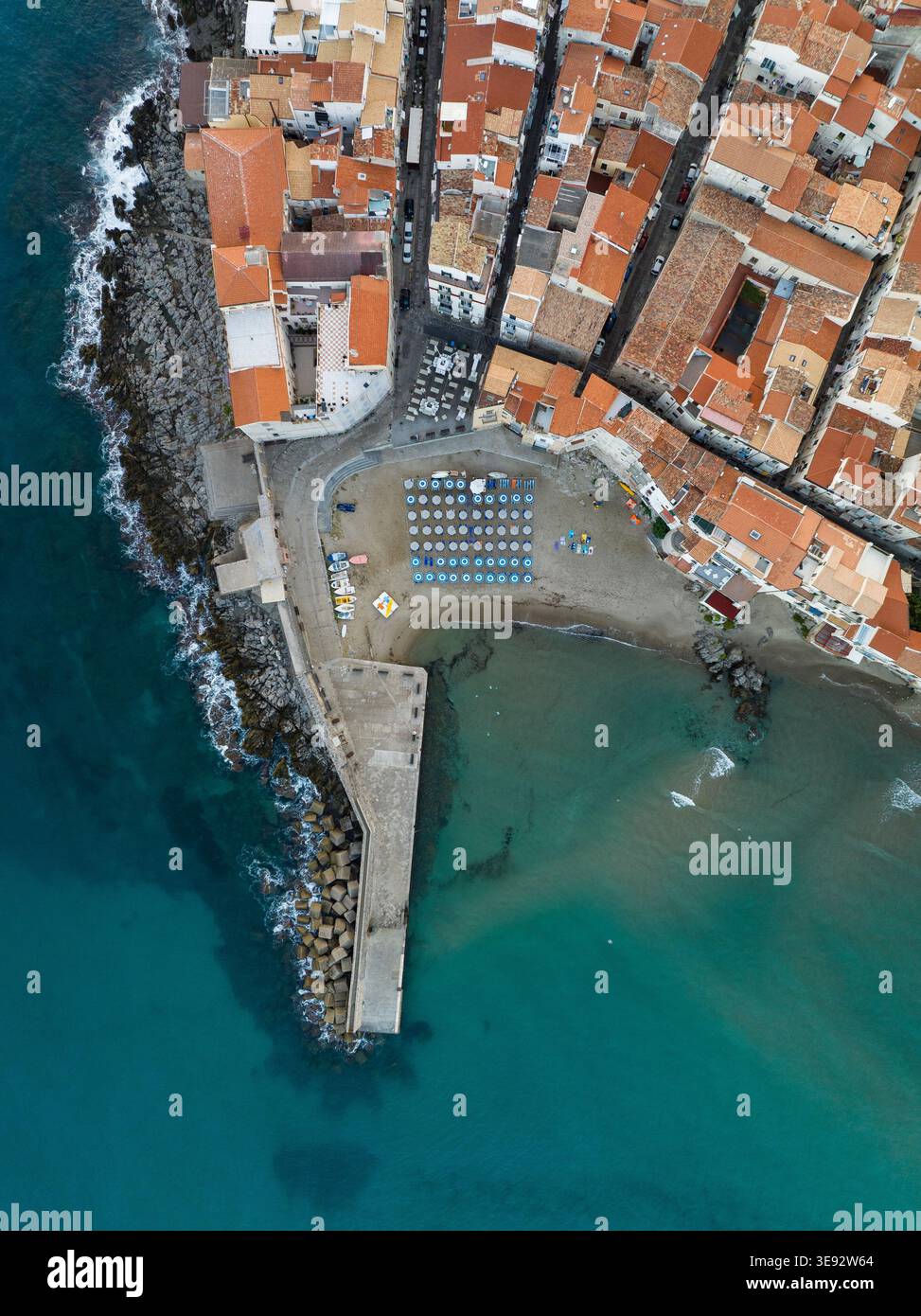 Veduta aerea di Cefalù con tetti arancioni, un molo in pietra e una spiaggia sabbiosa con ombrelloni blu lungo il mare turchese di Cefalù, Sicilia, Italia. Foto Stock