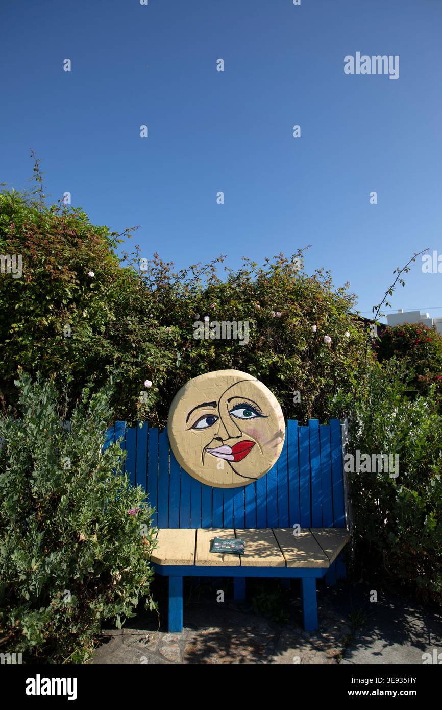 Half Moon Bay, California, USA - 31 ottobre 2025: Il sole pomeridiano illumina una panchina della città con una luna felice, circondata da piante. Foto Stock