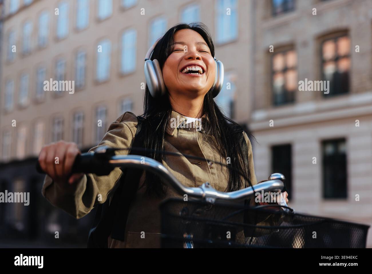 Una giovane donna ride mentre corre in bicicletta lungo una strada urbana, mette le mani sul manubrio e indossa cuffie sovrauricolari, ascoltando musica durante un su Foto Stock