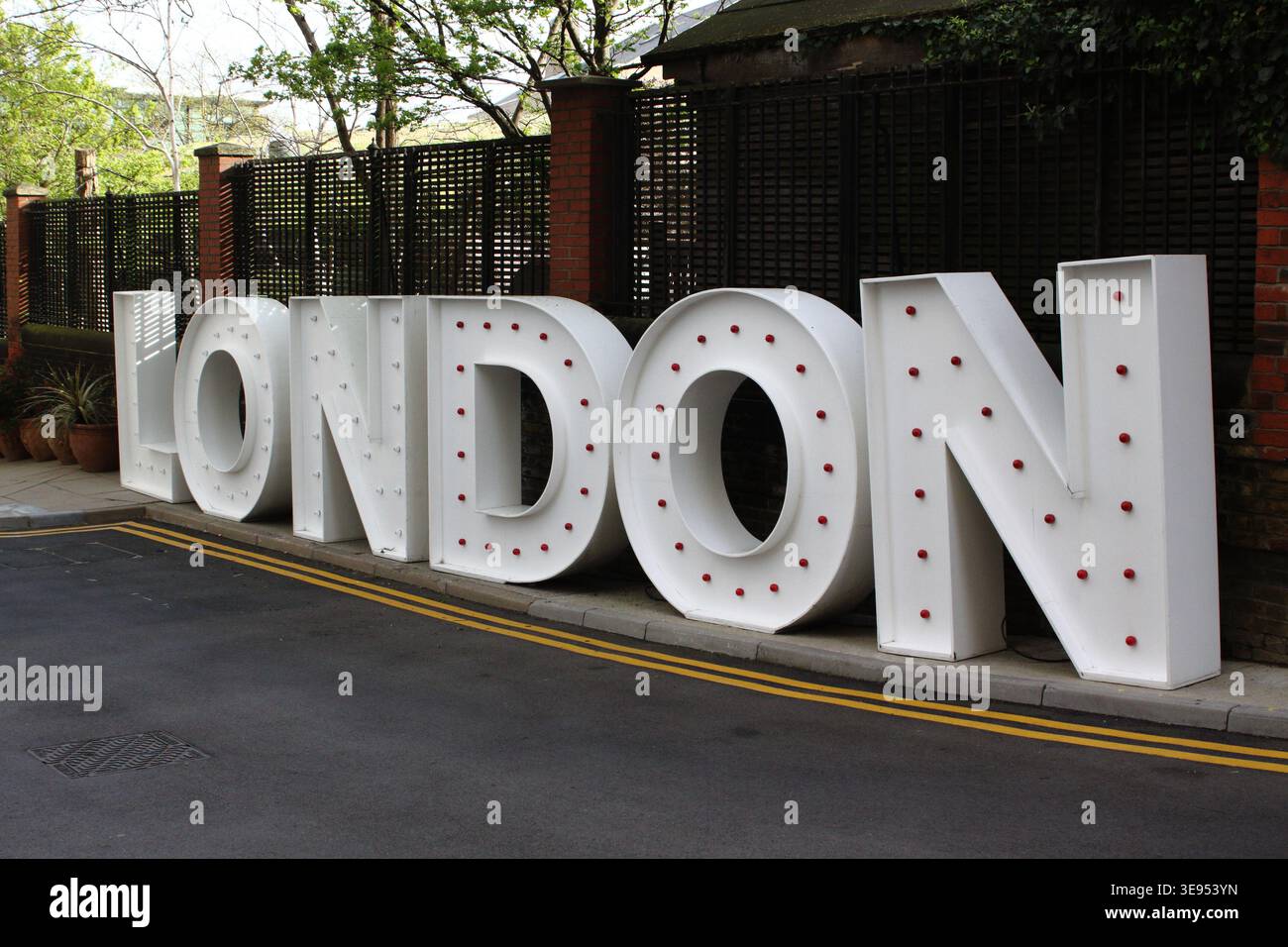 Grandi lettere bianche con tendone che scrivono londra su una strada cittadina accanto a muro e recinzione di mattoni, scena urbana all'aperto con alberi, di giorno. Foto Stock
